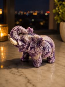 Natural hand-carved Amethyst crystal elephant statue with an upward-raised trunk, polished purple gemstone finish, displayed on a marble surface with a soft blurred background.